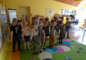 Grupa dzieci stoi przed ekranem z jedną ręką uniesioną a drugą skierowaną w dół, uczestniczą w zabawie ruchowej.